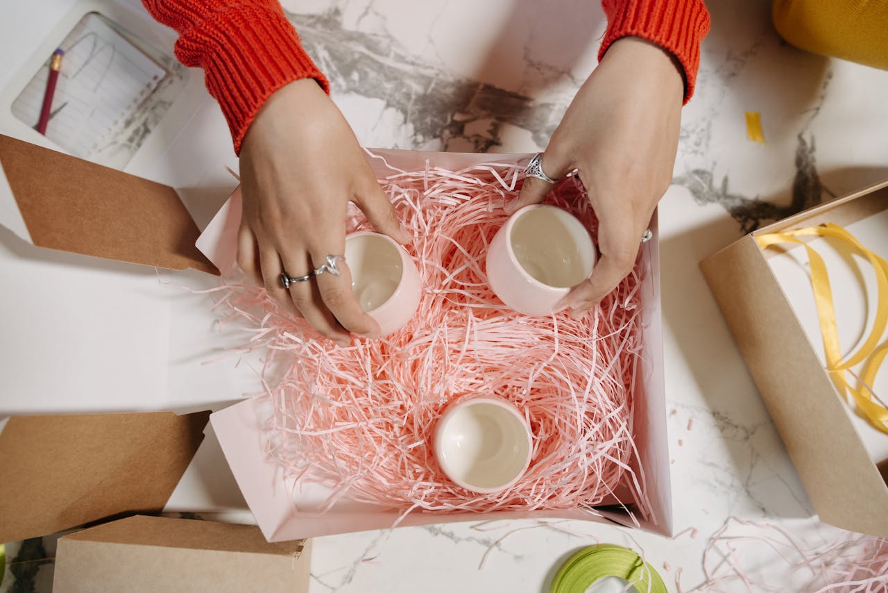Top view of hands arranging ceramic cups in a gift box with pink shredded paper.